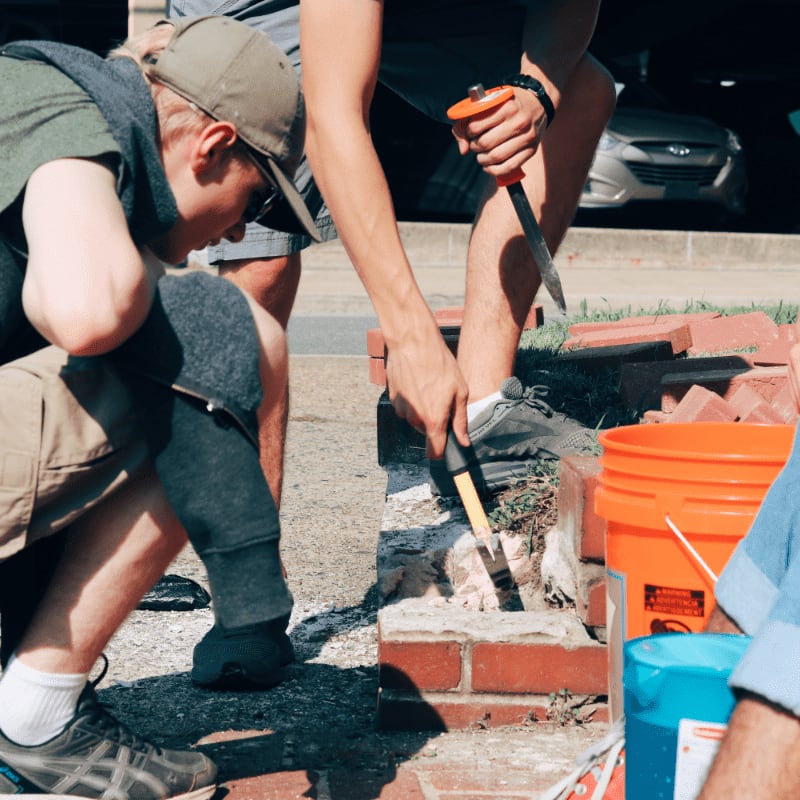 Two young men in the foreground are working on a brick structure with tools and buckets, while a car is visible in the blurred background.