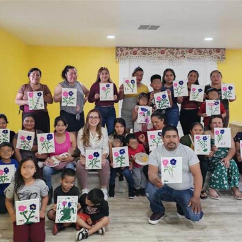 A group of adults and children proudly display their colorful flower paintings in a brightly lit room with yellow walls.