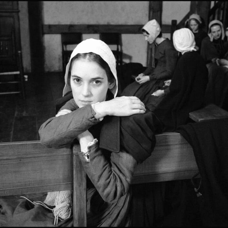 A young woman in a bonnet and period clothing rests her arms on a wooden pew in the foreground, while other women in similar attire sit behind her in a dimly lit room.