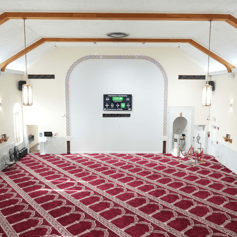 A spacious prayer hall with a red patterned carpet in the foreground and a large digital display on a white wall in the background.