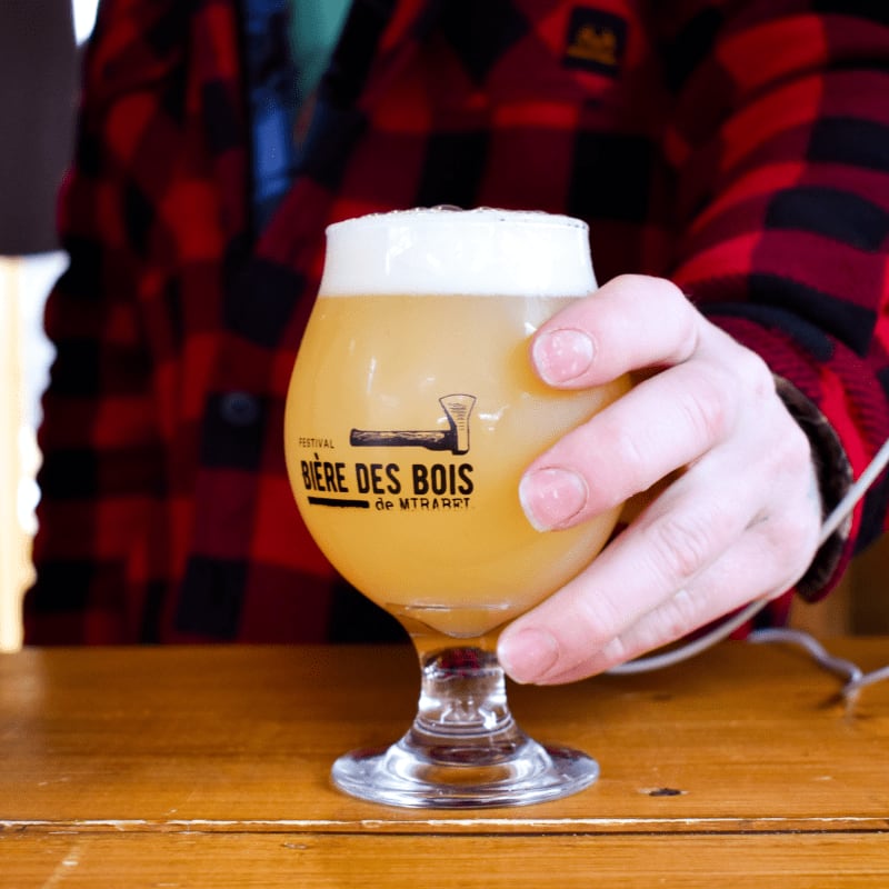 A hand in a red plaid shirt reaches for a glass of frothy beer with a logo on a wooden table.