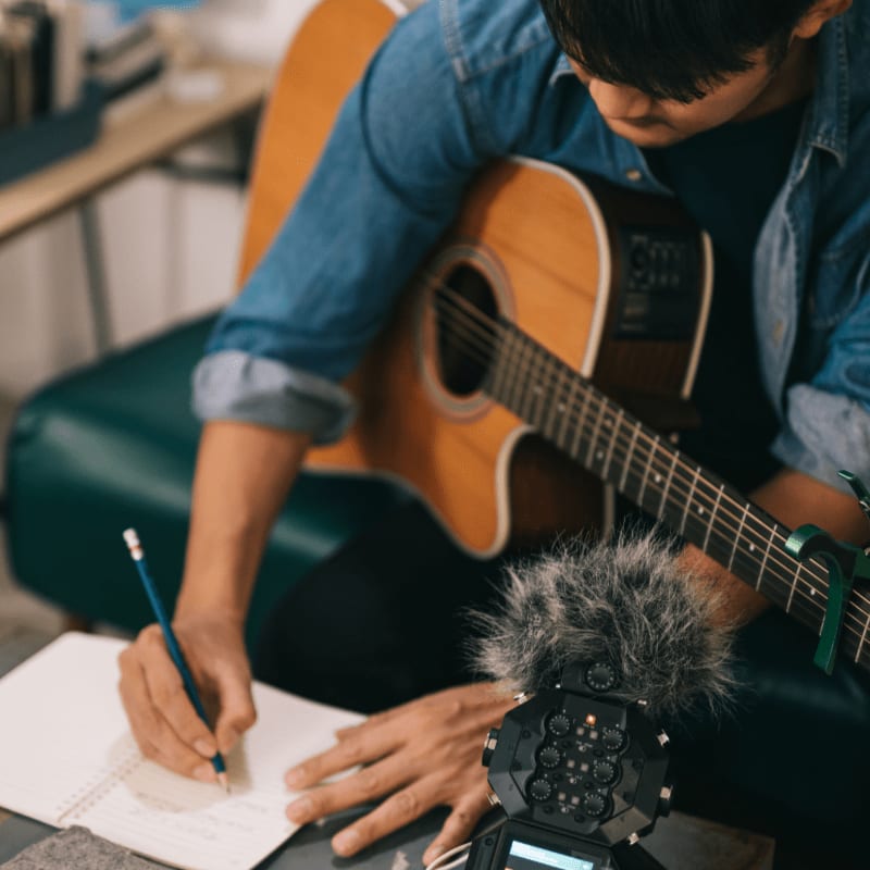 A person in a denim shirt plays an acoustic guitar while writing in a notebook, with a microphone and recording device in the foreground and a blurred background.