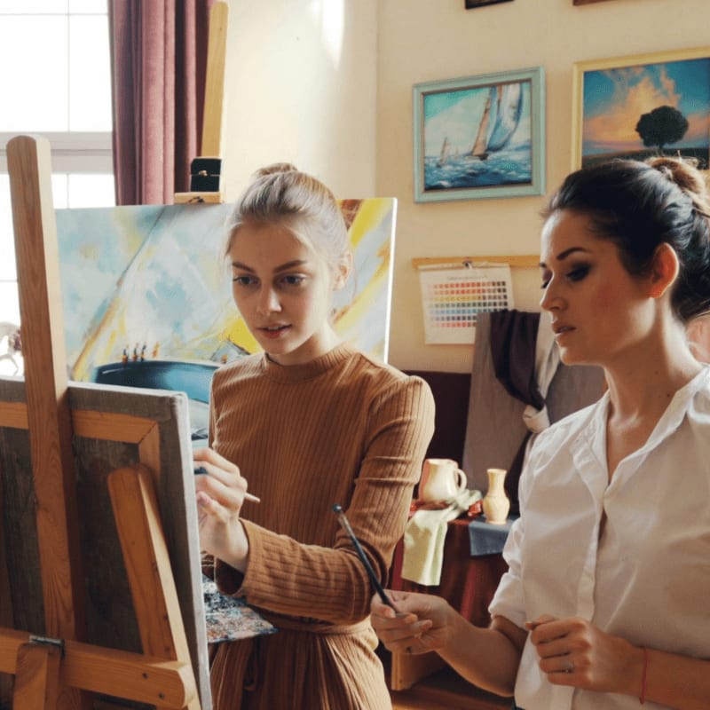 Two women are painting in an art studio, with canvases and framed artwork visible in the background.