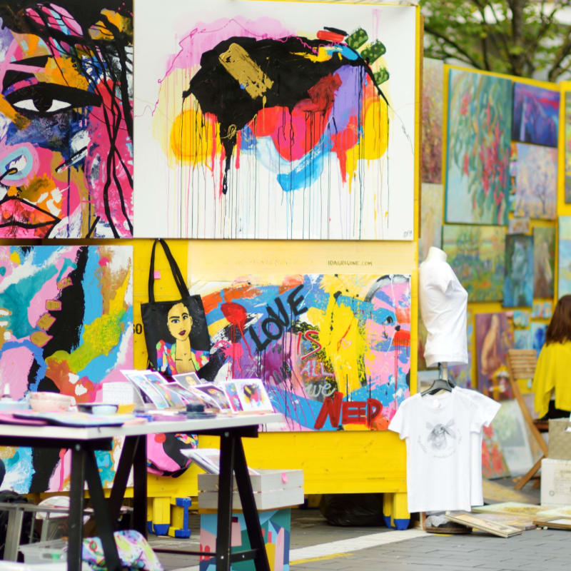 Colorful abstract paintings are displayed on yellow stands in the background, with a table of smaller artworks and merchandise in the foreground.