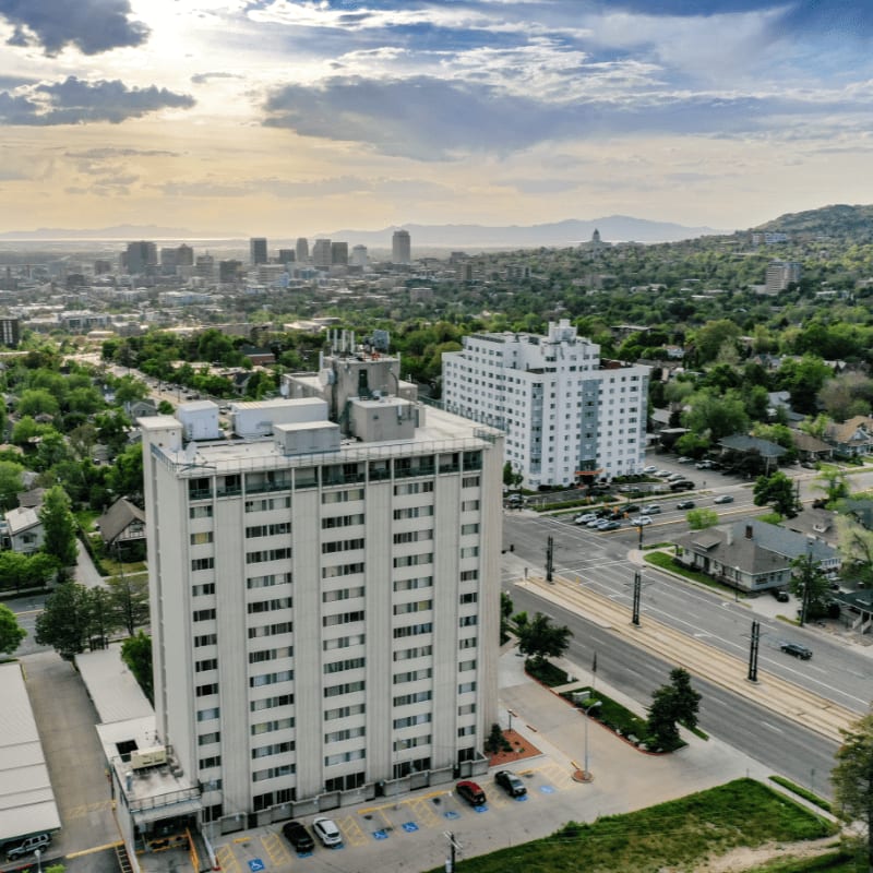 A tall, white apartment building with many windows dominates the foreground, while a sprawling city with distant mountains and a cloudy sky forms the background.