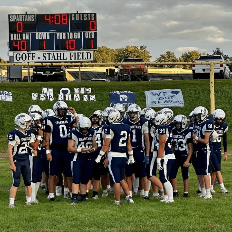 A football team in navy and white uniforms huddles on a grassy field in front of a scoreboard displaying "SPARTANS 0" and "GUESTS 0" with a line of vehicles and trees in the background.