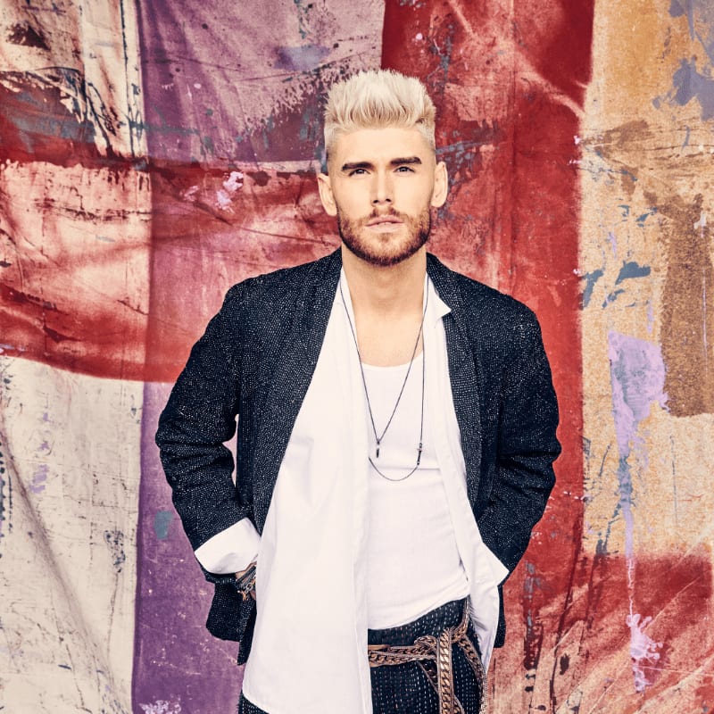 A man with blonde, spiky hair and a beard stands in the foreground wearing a dark jacket over a white shirt and a decorative belt, against a colorful, abstract background of red, purple, and white fabric.
