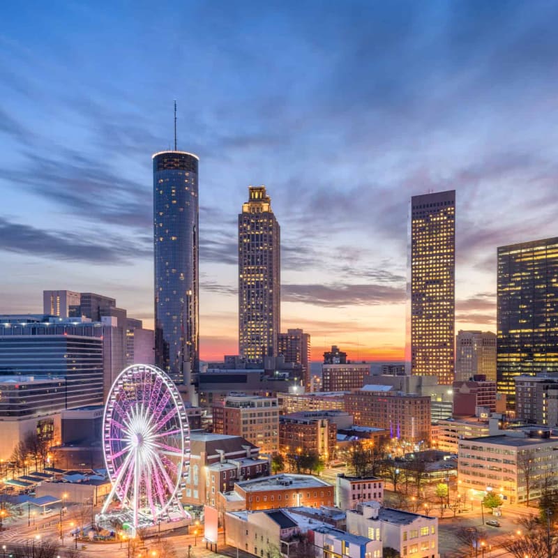 A brightly lit Ferris wheel stands in the foreground of a city skyline at dusk, with tall buildings illuminated against a colorful sky.