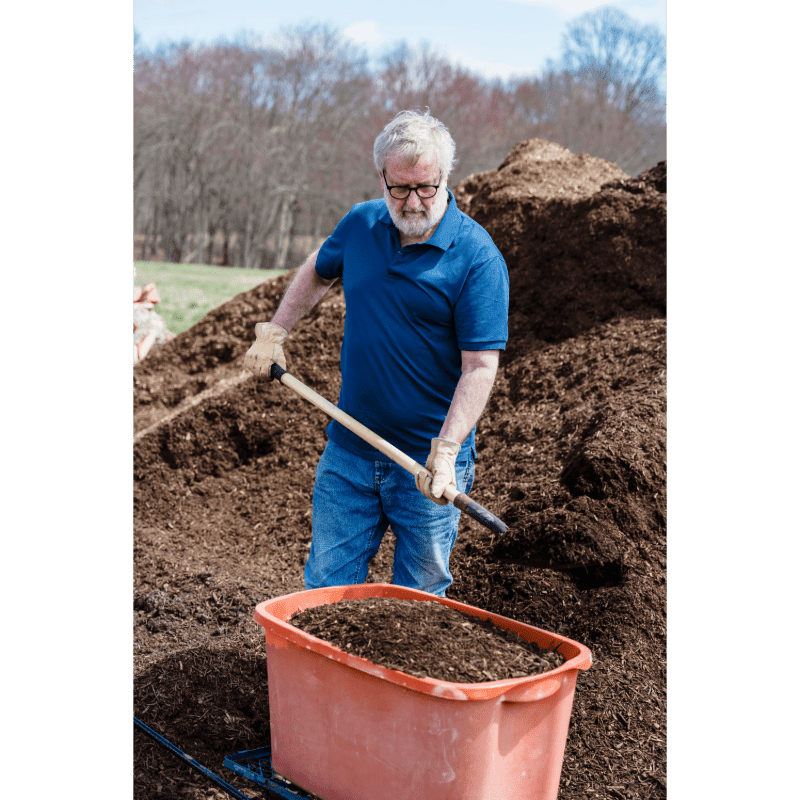 A man in a blue shirt and jeans shovels mulch into an orange wheelbarrow in front of large piles of mulch with trees in the background.