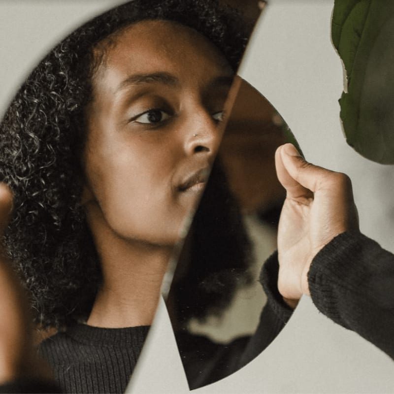 A woman with curly hair looks into a fragmented mirror, her reflection partially obscured by a hand in the foreground against a soft, neutral background.