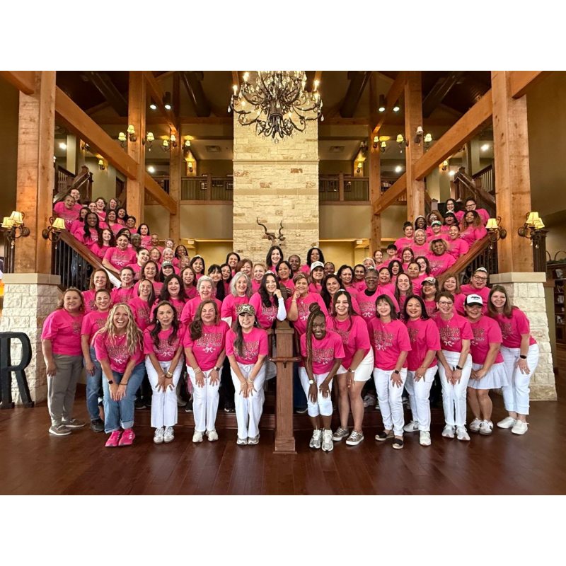 A large group of women in pink shirts and white pants pose on a wooden staircase with a stone accent wall and a chandelier in the background.