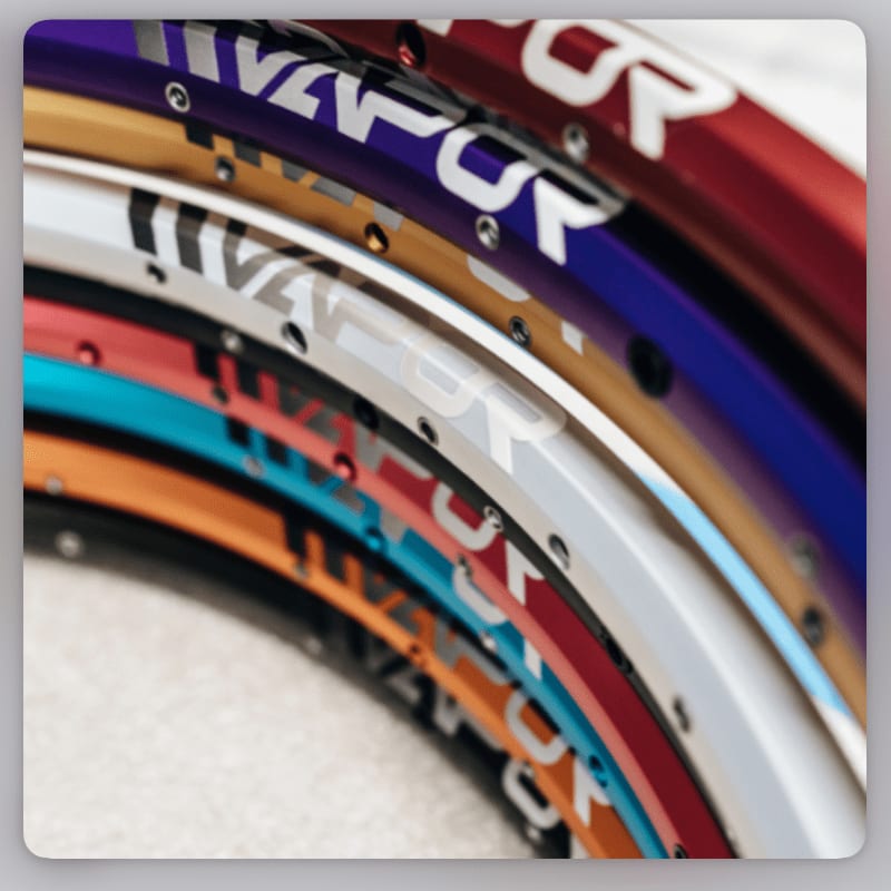 A stack of colorful bicycle rims with the "VAPOR" logo is in the foreground, against a blurred, light-colored background.