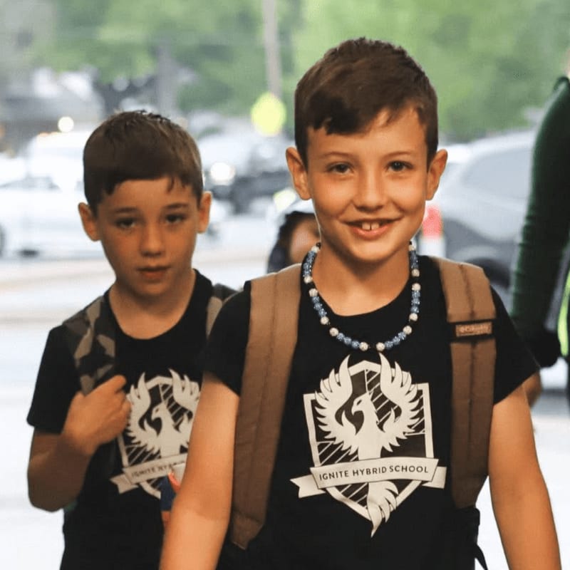 Two young boys wearing matching t-shirts and backpacks walk forward, with blurred cars and buildings in the background.