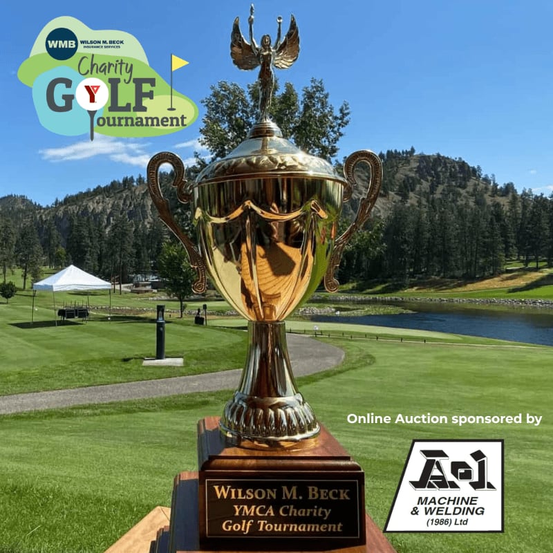 A large golden trophy stands prominently in the foreground on a wooden base, with a scenic golf course, lake, and tree-covered hills forming the background under a clear blue sky.