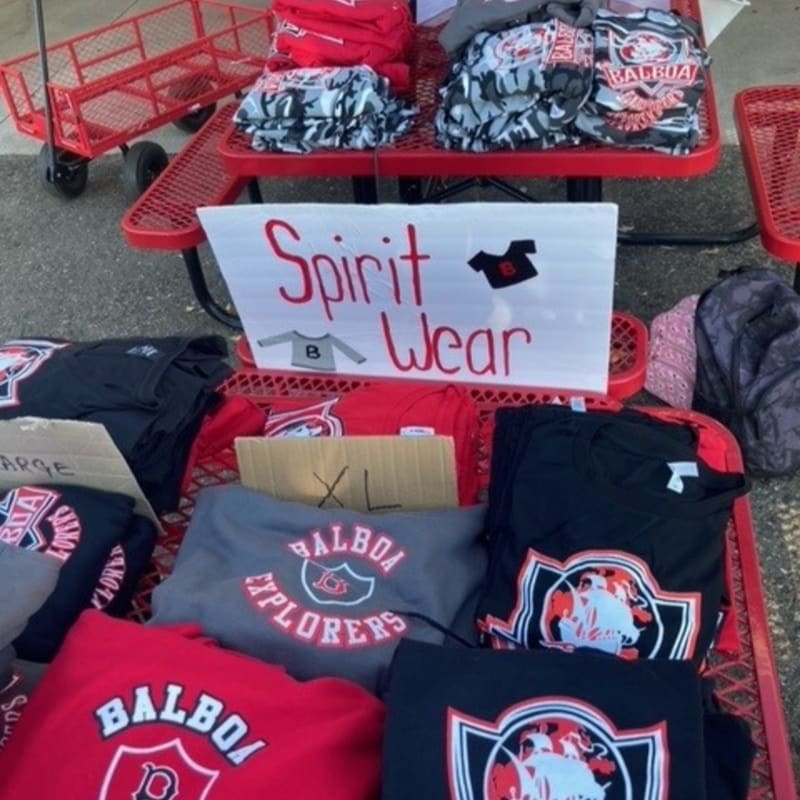 Red spirit wear is displayed on a red picnic table in the foreground, with a red wagon and more merchandise visible in the background.