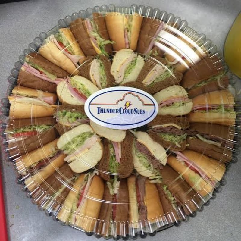 A circular platter of assorted mini sandwiches is arranged on a gray surface, with a yellow drink visible in the upper right corner.