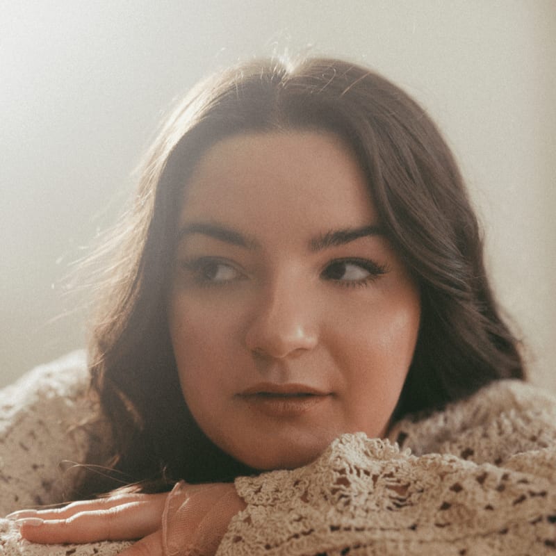 A young woman with dark hair rests her head on her arms, looking away from the camera against a softly lit, neutral background.