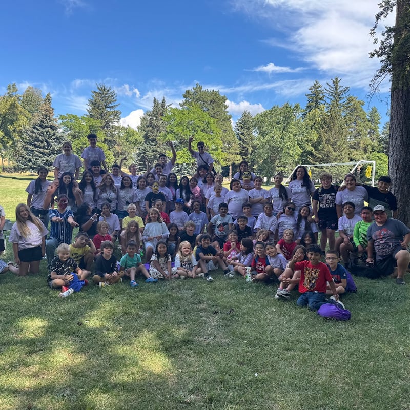 A large group of people, mostly children and young adults, are gathered on a grassy lawn in the foreground, with a backdrop of trees and a bright blue sky with scattered clouds.