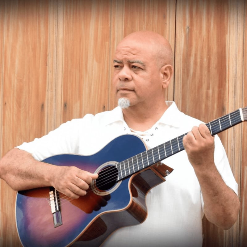 A bald man with a goatee plays an acoustic guitar in front of a wooden wall.