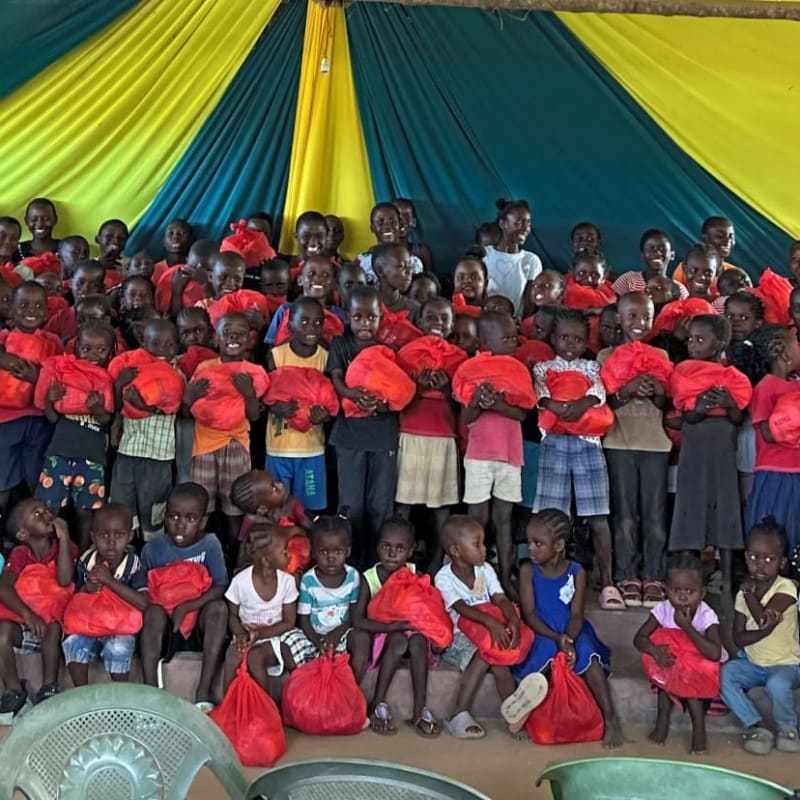 A large group of children in the foreground, many holding red bags, are gathered under a yellow and green striped tent in the background.