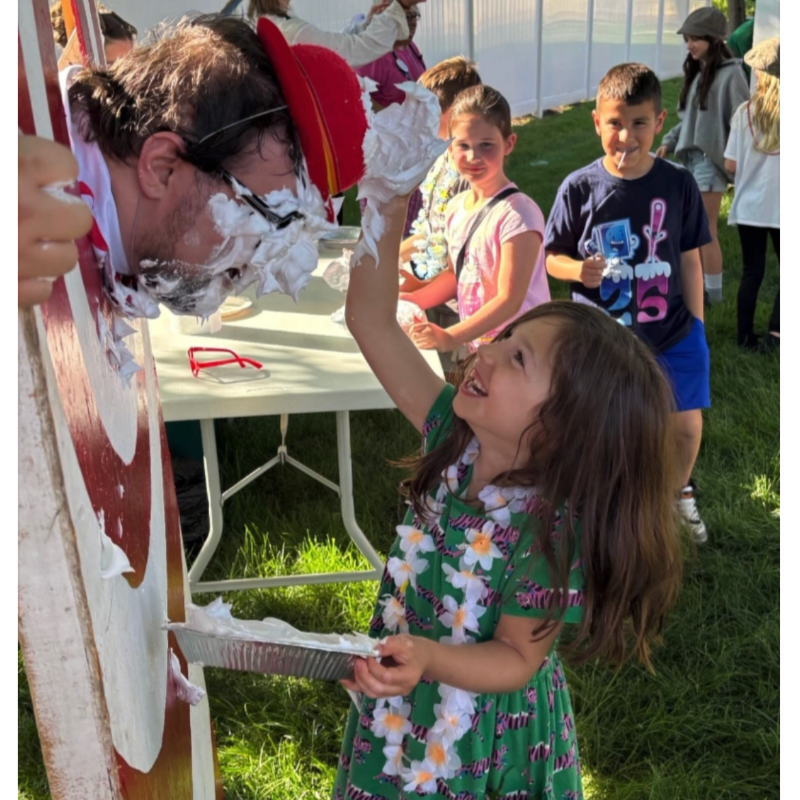 A young girl in a green floral dress throws a pie at a man's face in the foreground, while other children watch in the background.