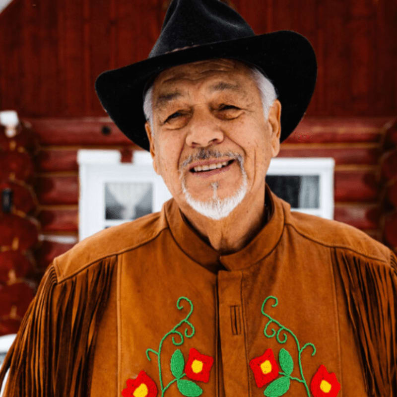 A smiling Indigenous man in a beaded jacket and black hat stands before a rustic log cabin.