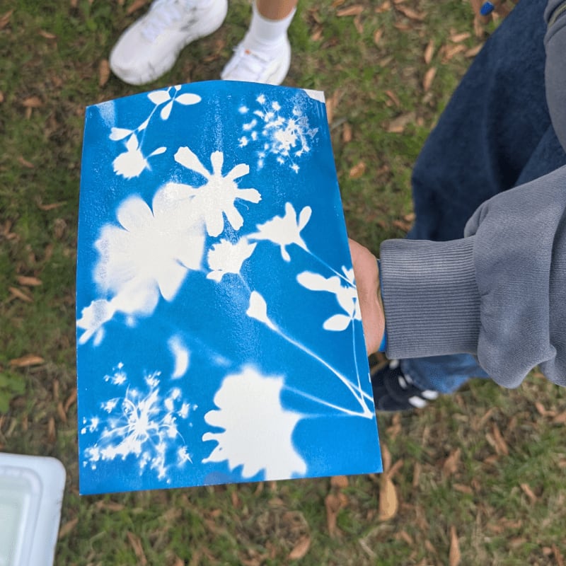 A hand holds a blue and white floral-patterned object in the foreground, with people's legs and grassy ground in the background.