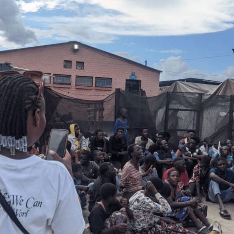 A woman in the foreground films a crowd gathered outside a pink building under a cloudy sky.