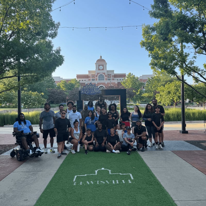 A diverse group of people, including someone in a wheelchair, pose on a green turf with a building and trees in the background.