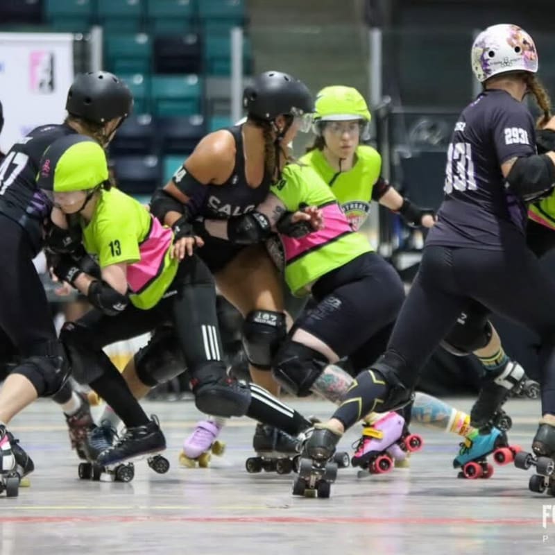 In the foreground, roller derby skaters in colorful uniforms jostle for position, while the background shows a blurred indoor arena.