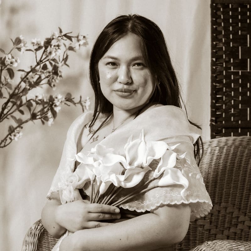 A woman in traditional attire sits in a wicker chair holding calla lilies, with a flowering branch to her left and a woven screen behind her.