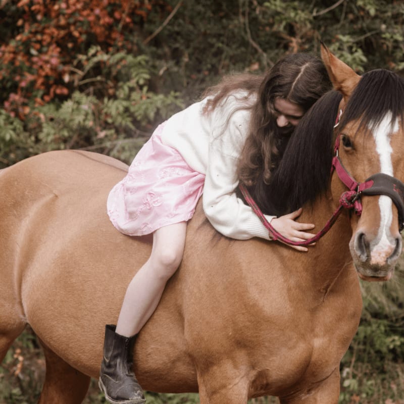 A young girl in a pink dress hugs a brown horse, with a blurred background of autumn foliage.
