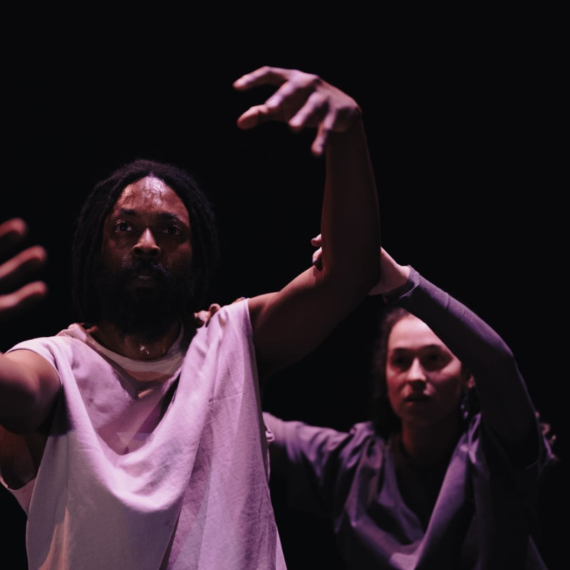 A man with dreadlocks in a white tunic and a woman in a grey top move their arms against a dark background.