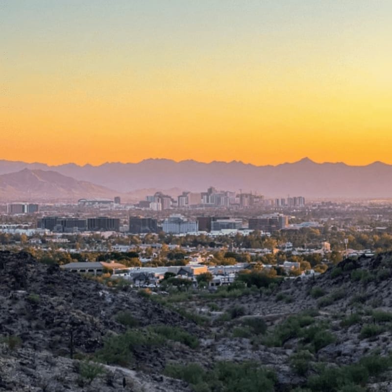 Rocky, scrub-covered hills in the foreground give way to a sprawling city nestled against a hazy mountain range under a warm, gradient sky.