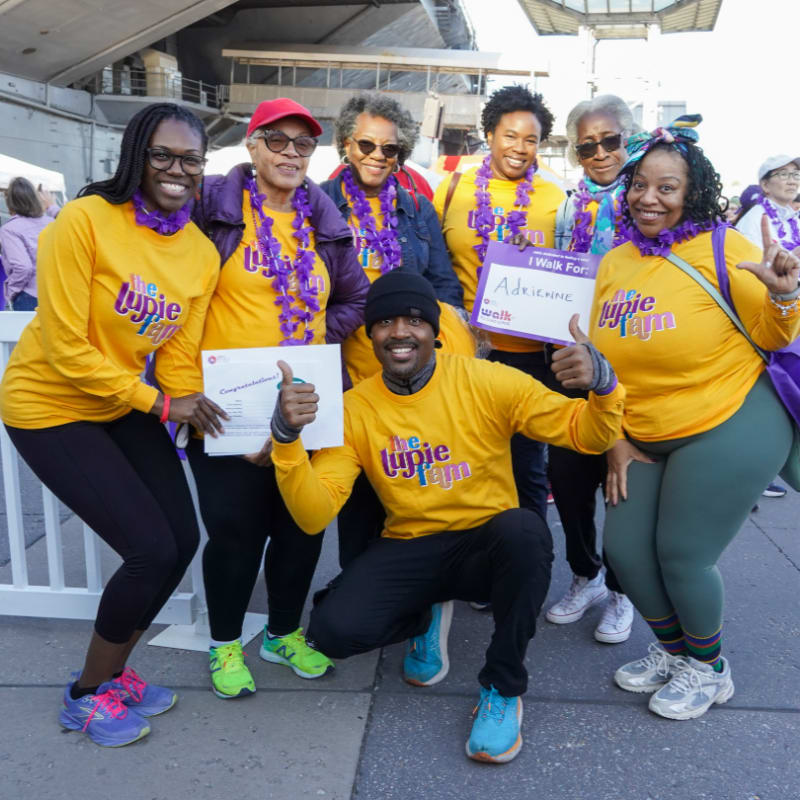 A group of people in yellow shirts and purple leis pose for a photo in front of a large building.