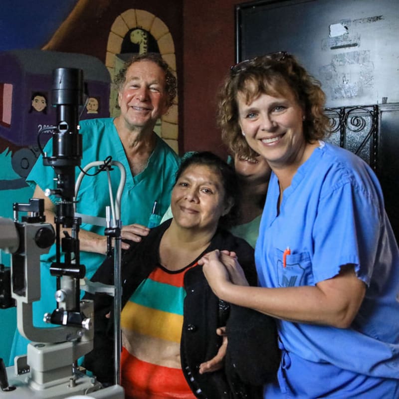 A smiling woman in colorful clothing is flanked by two medical professionals in scrubs, with an eye examination machine in the foreground and a mural of a train in the background.