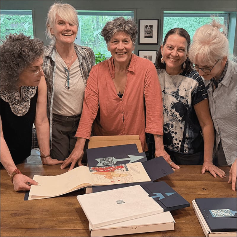 Five smiling women gather around a wooden table with open folders and books, while a blurred green background suggests an outdoor setting.