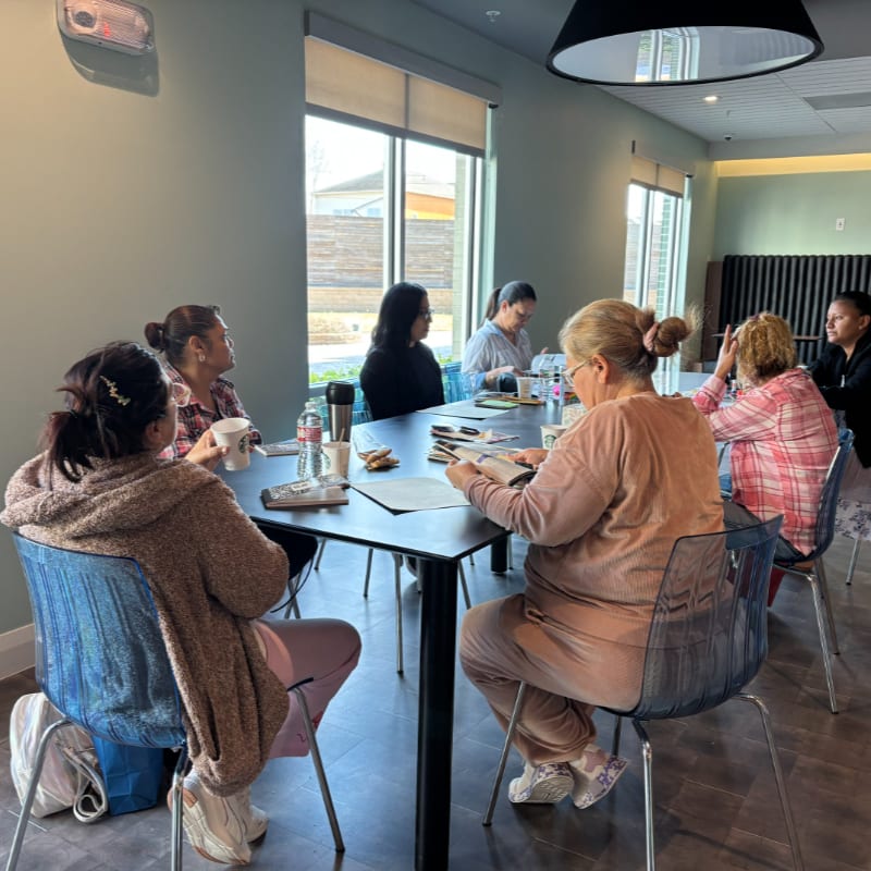 Several women sit around a table in a brightly lit room with large windows in the background.