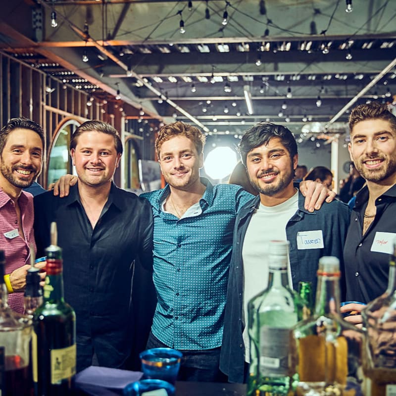 Five smiling men pose for a photo in the foreground, with bottles of alcohol on a bar, while a dimly lit event space with string lights is visible in the background.