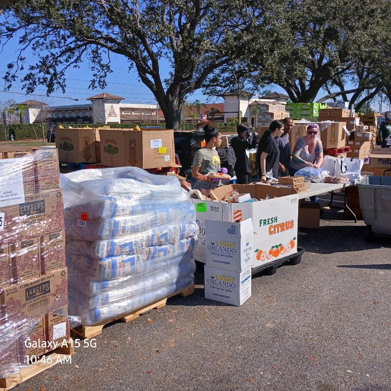 In the foreground, stacks of boxed goods wrapped in plastic are visible, while in the background, people are gathered around tables laden with food, with trees and buildings behind them.