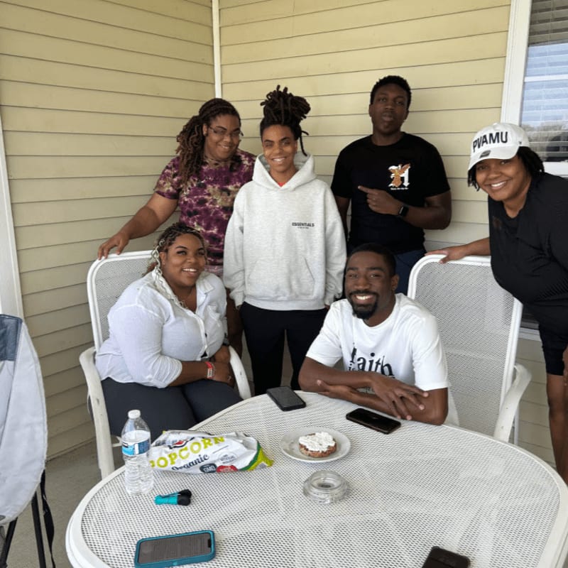 Six smiling people pose for a photo on a porch with a table in the foreground holding snacks and drinks.
