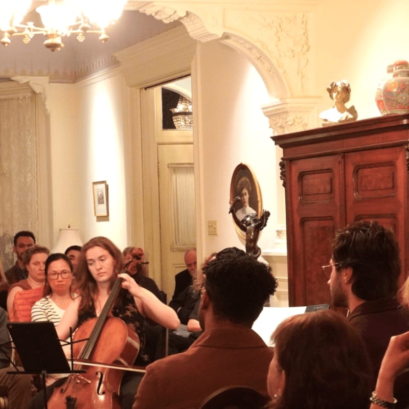 A woman plays the cello in the foreground while an audience watches in a richly decorated room with a large wooden cabinet in the background.