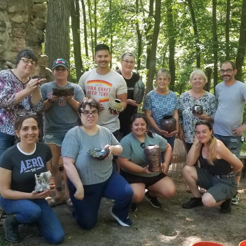 A group of people pose with their pottery in a sun-dappled forest clearing.
