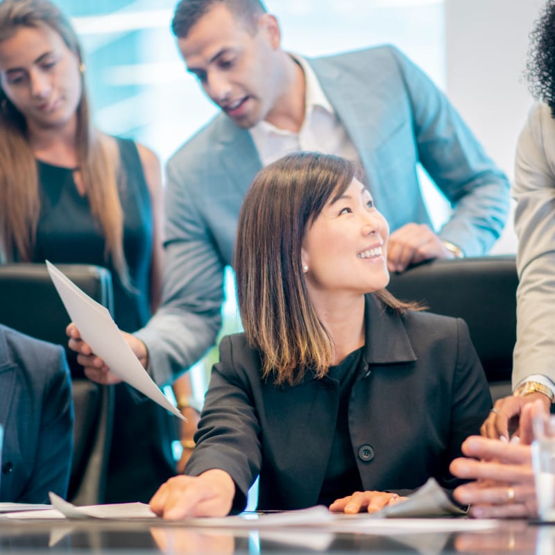 A smiling Asian woman in a black suit is in the foreground, while a diverse group of colleagues in business attire are gathered around a table in the background.