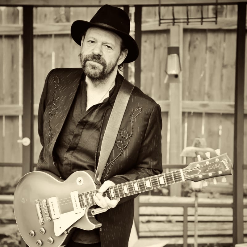 A man in a hat and suit holds a guitar in front of a wooden fence.