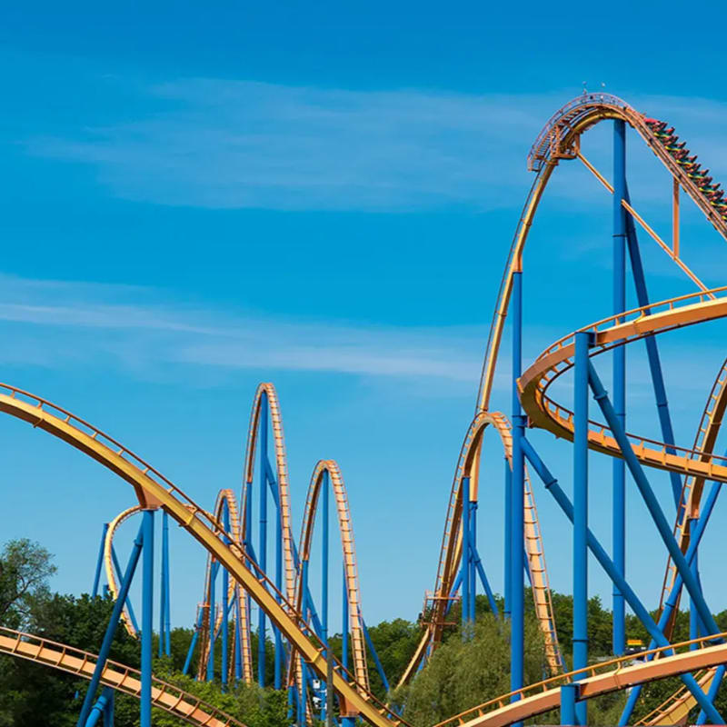 A bright blue sky serves as the backdrop for a towering orange and blue roller coaster with lush green trees visible below.