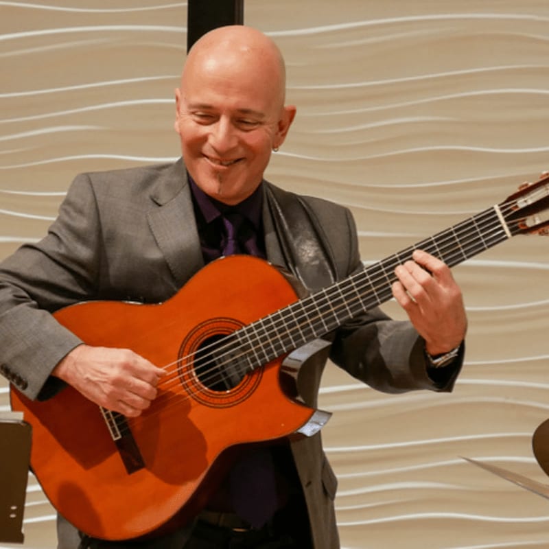 A bald man in a suit plays a guitar in front of a textured, wavy wall.