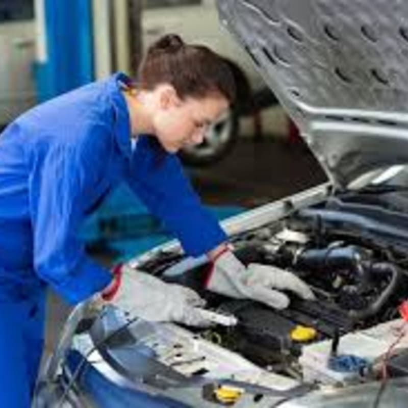 A mechanic in blue overalls and gloves works on the engine of a car with the hood open, while a blurred background suggests a garage setting.