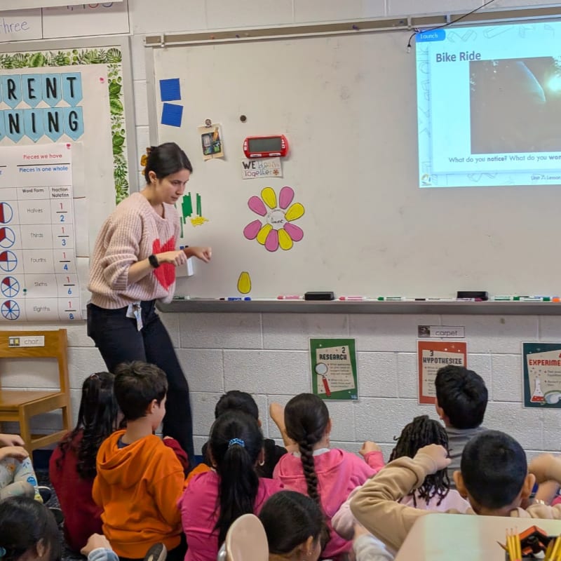 A teacher stands in front of a whiteboard with students sitting on the floor in the foreground, while a presentation titled "Bike Ride" is displayed on the screen in the background.