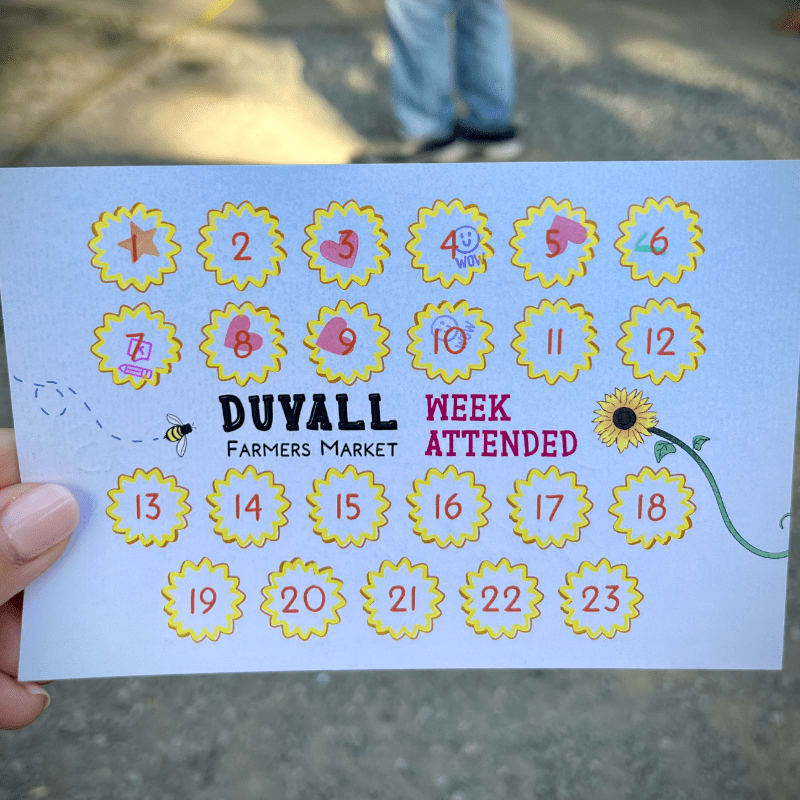 A hand holds a "Duvall Farmers Market Week Attended" card with numbered circles, while a person's legs and feet are visible in the blurred background.